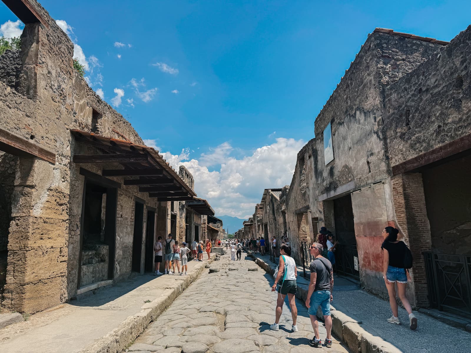 Tourists walking along an ancient stone street in Pompeii, with ruined Roman buildings on both sides under a bright blue sky.