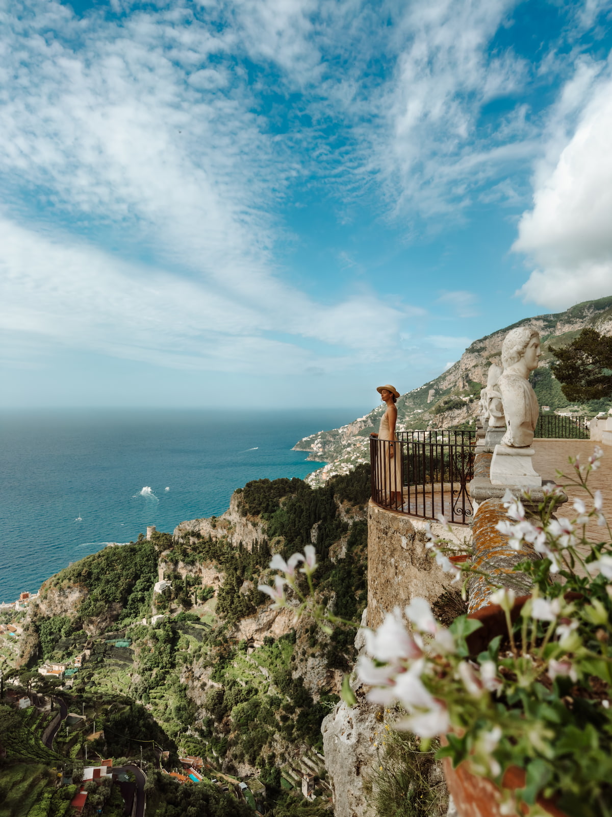 Woman in a straw hat standing on the Terrace of Infinity at Villa Cimbrone in Ravello, with marble busts, flowers, and sweeping Amalfi Coast sea views.