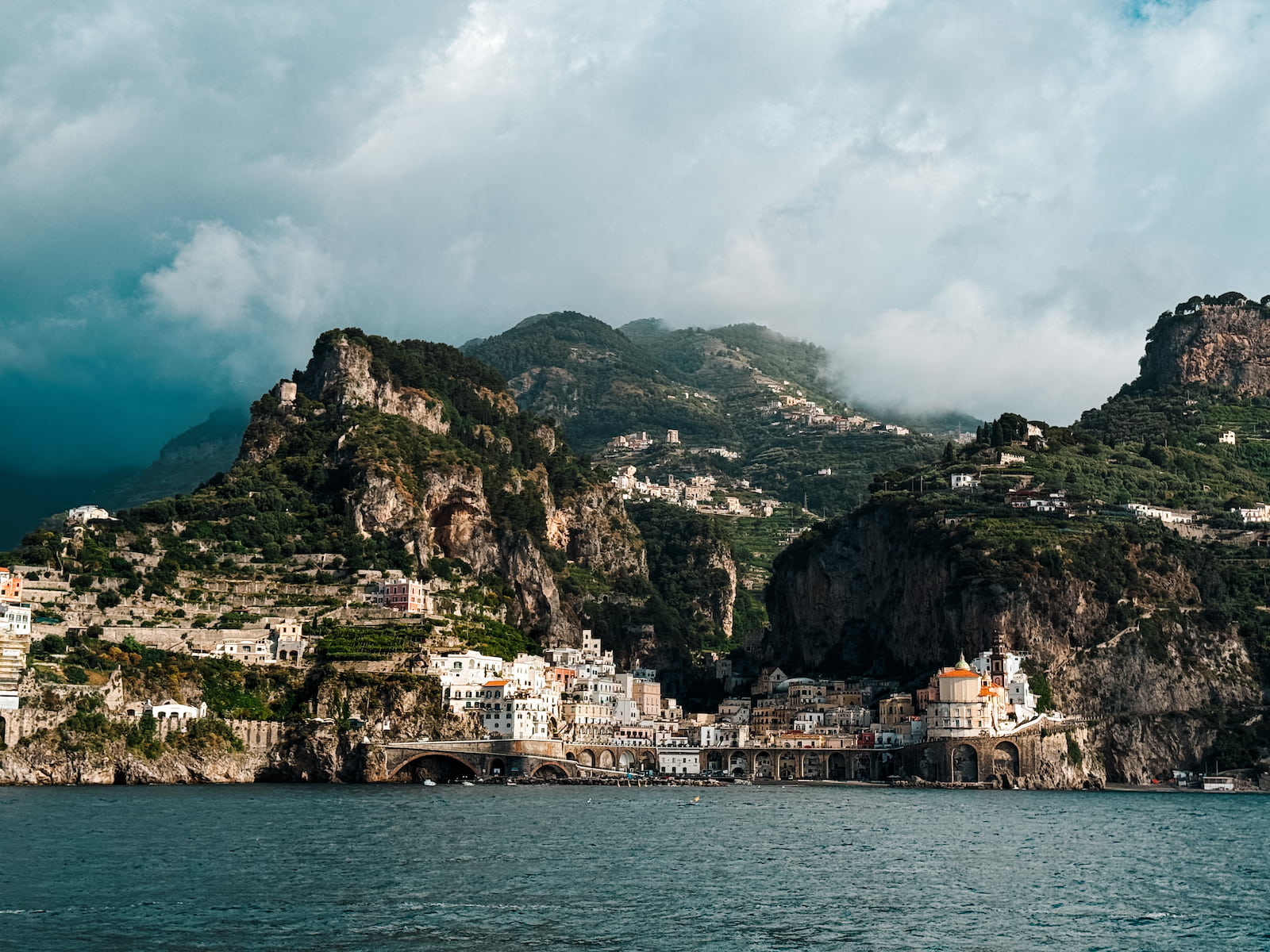 View of Atrani from the water, with the Amalfi Coast village framed by steep cliffs, green hills, and low clouds.