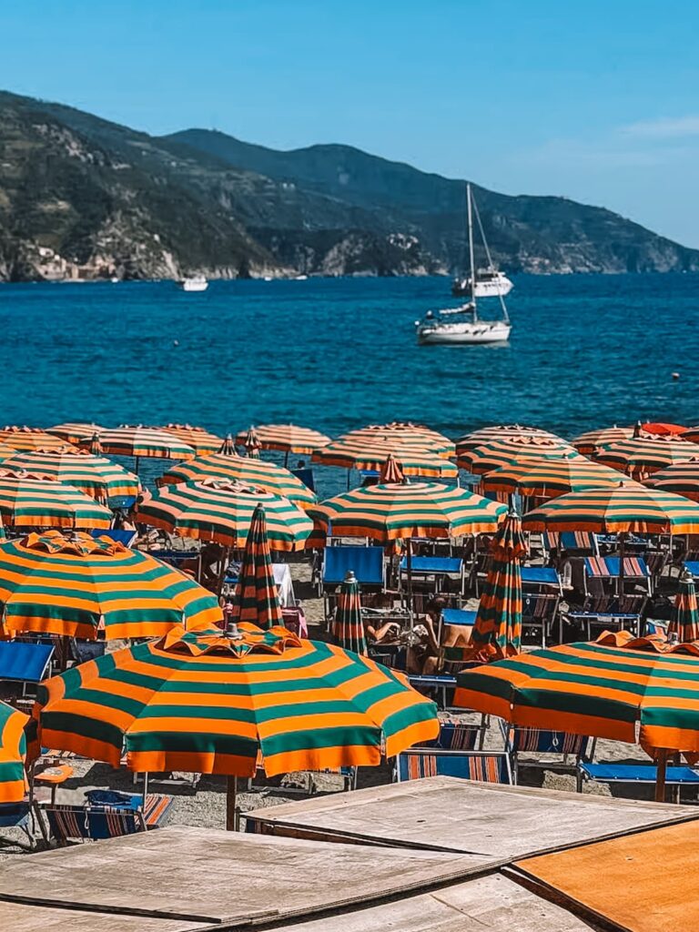 Beach umbrellas and lounge chairs on Monterosso beach, with sailboats and the Ligurian coastline in the background.