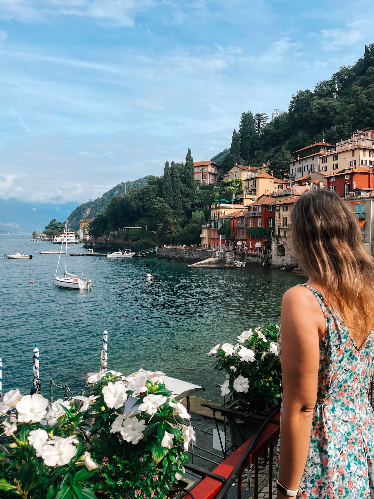 Woman overlooking Lake Como from a balcony in Varenna, with colorful waterfront houses, boats, flowers, and mountains in the background.
