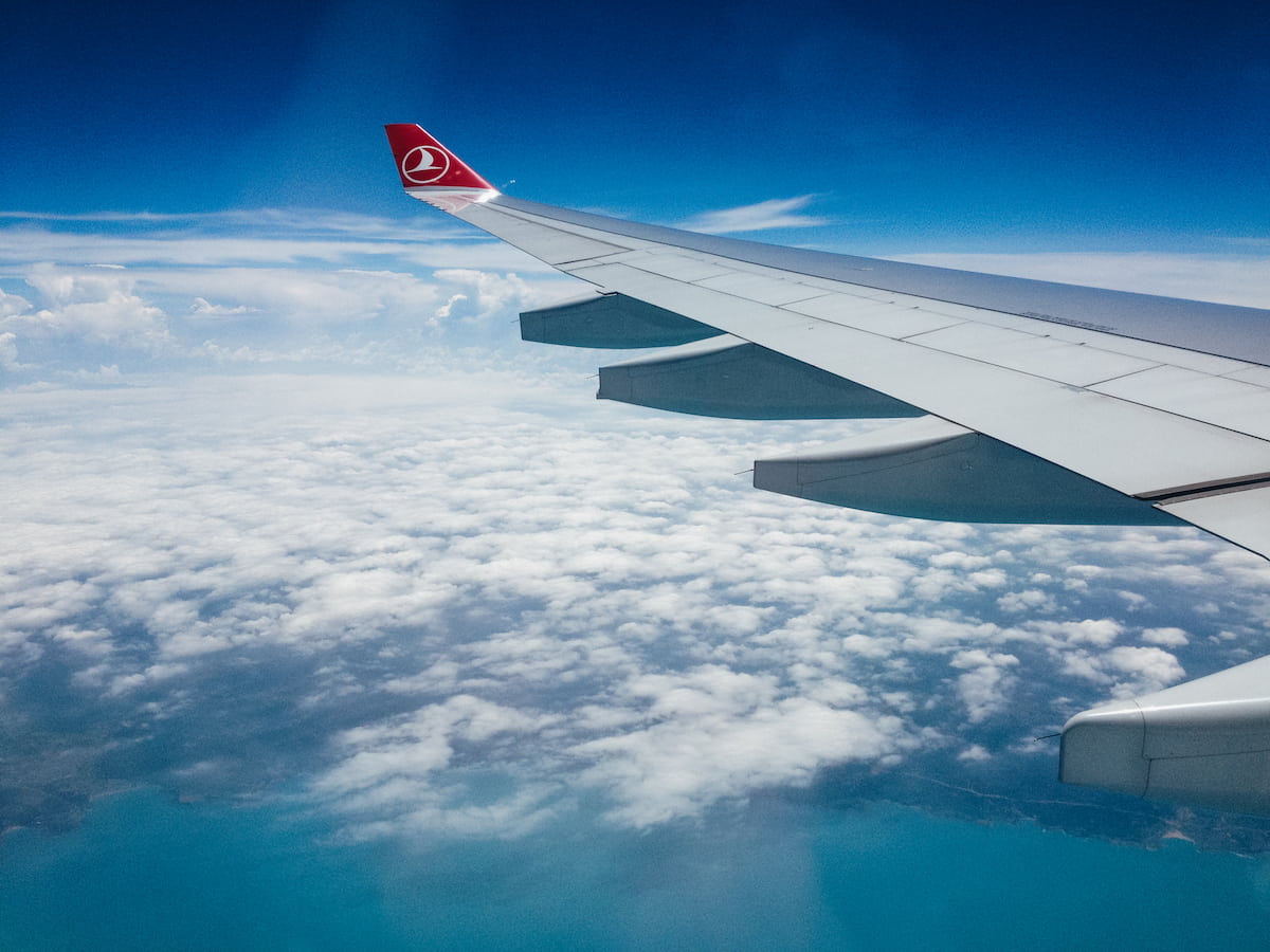 Turkish Airlines airplane wing above clouds and a turquoise coastline, viewed from a passenger window.